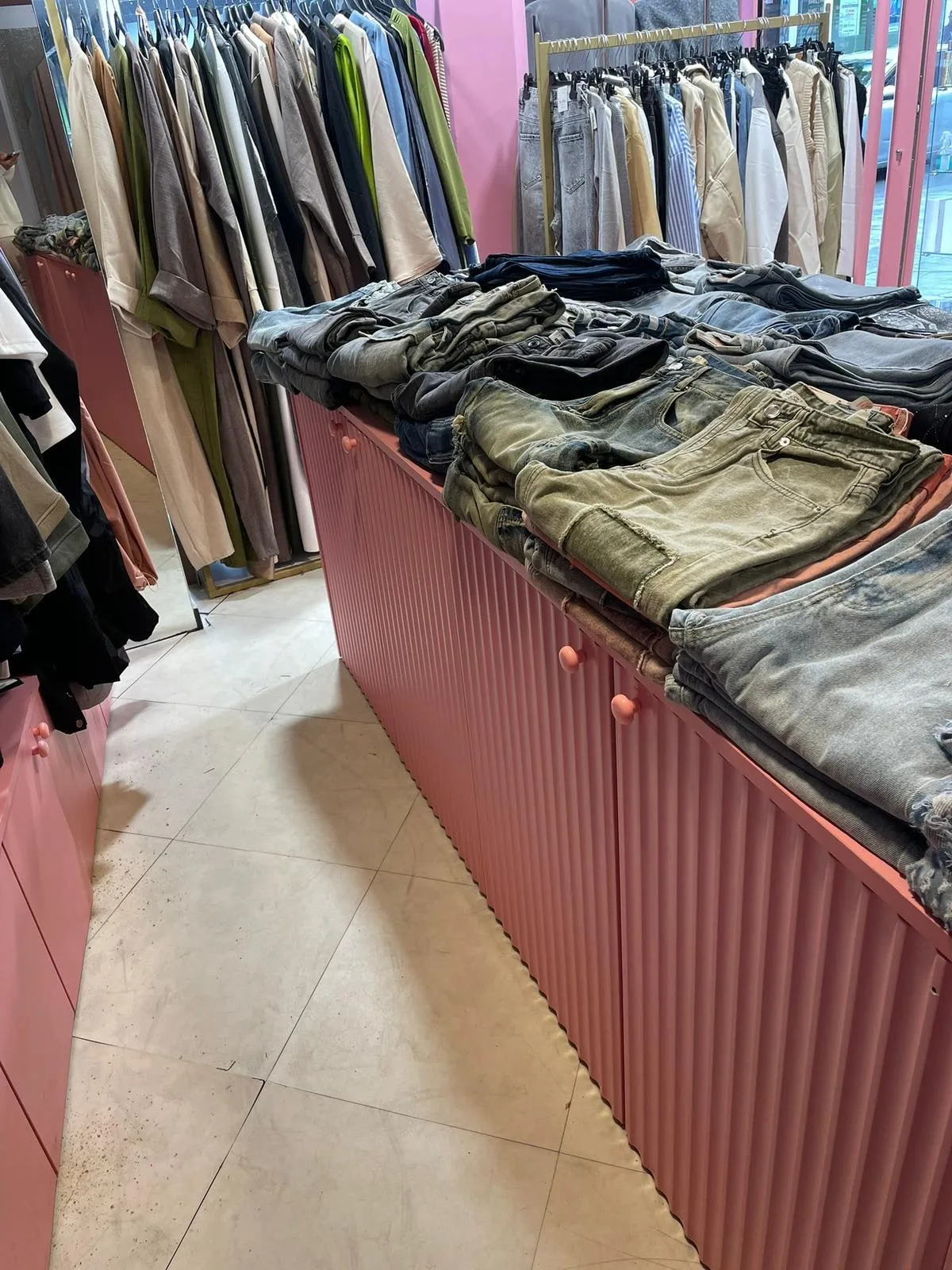 Meuble de présentation rose a façade cannelée avec jeans plies, portants dores et miroir en boutique