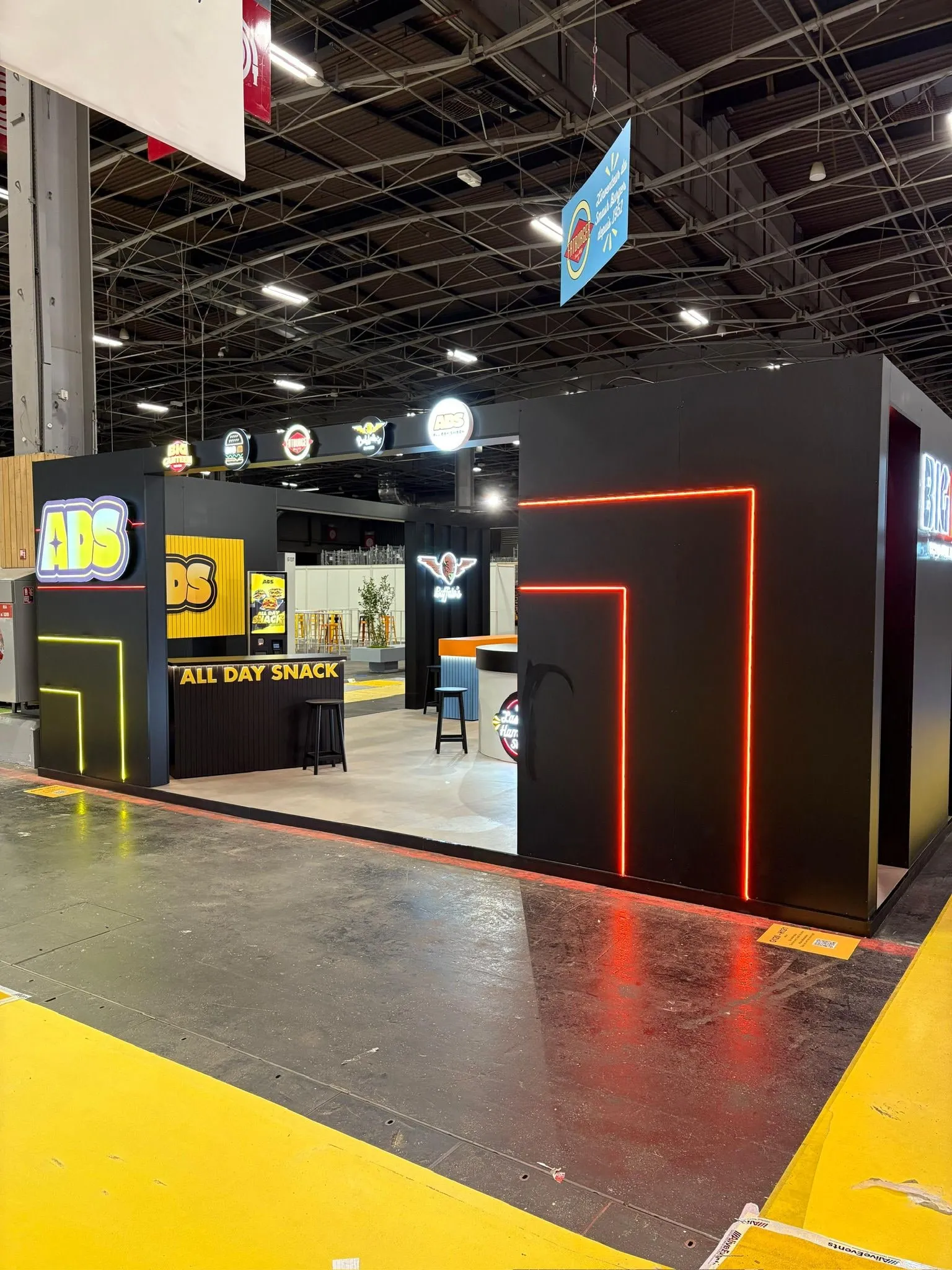 Façade du stand : panneaux noirs a neons lineaires jaune et rouge, comptoir a tasseaux en retrait