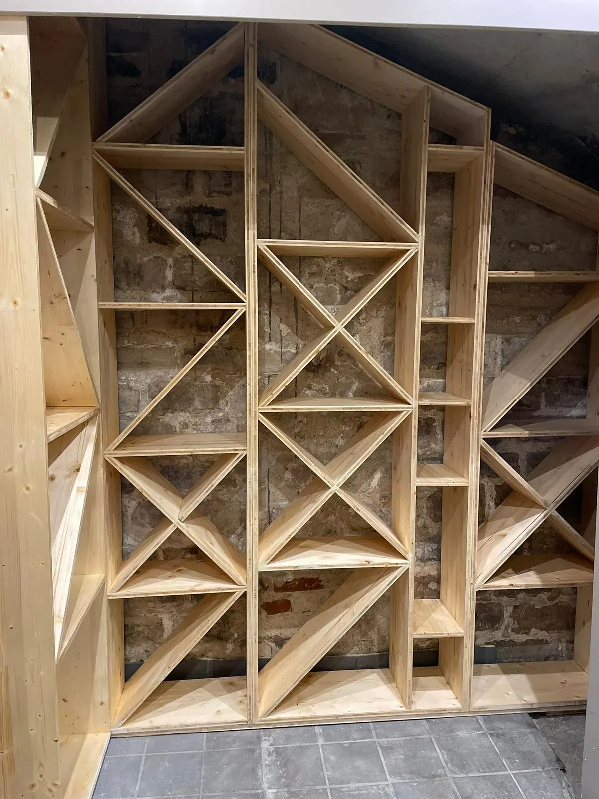 Casier a bouteilles en bois massif a cases triangulaires et croisillons, adosse a un mur en pierre apparente