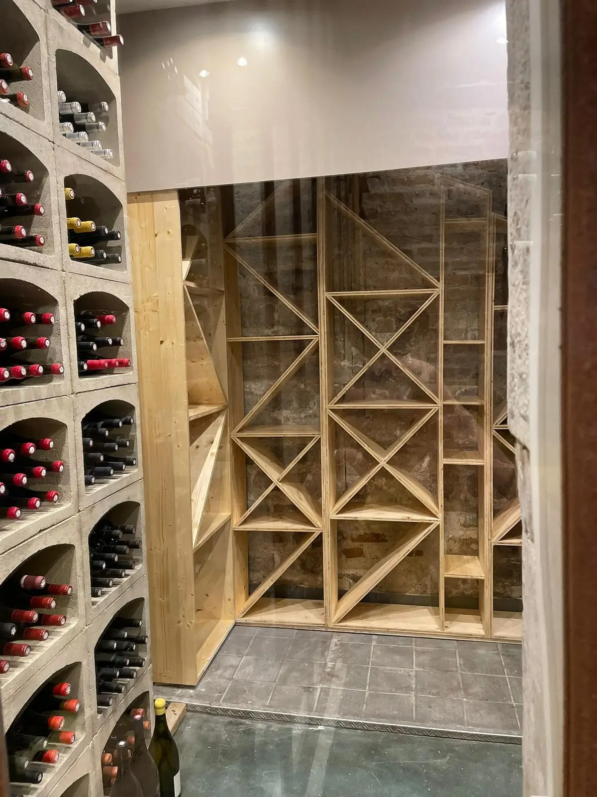 Cave à vin avec casier bois à croisillons et casiers béton remplis de bouteilles, vue depuis la porte vitrée