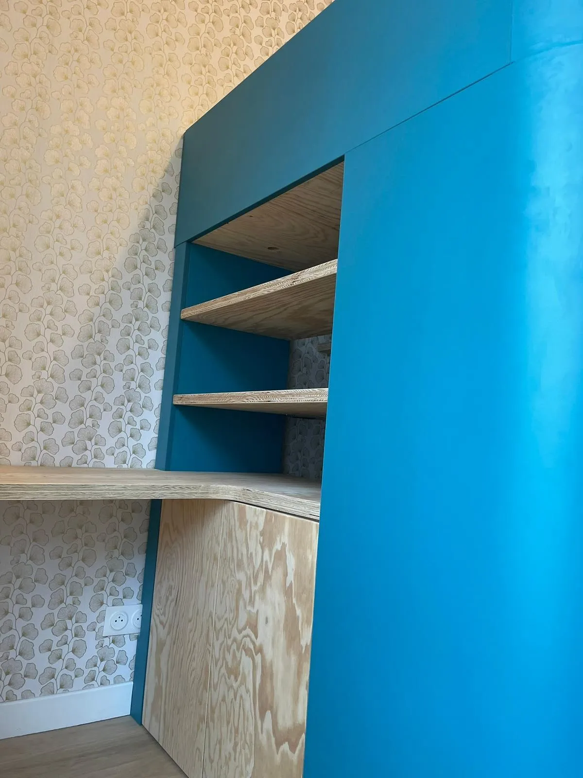 Bureau intégré en contreplaque sous une structure bleu canard, étagères et papier peint a feuilles