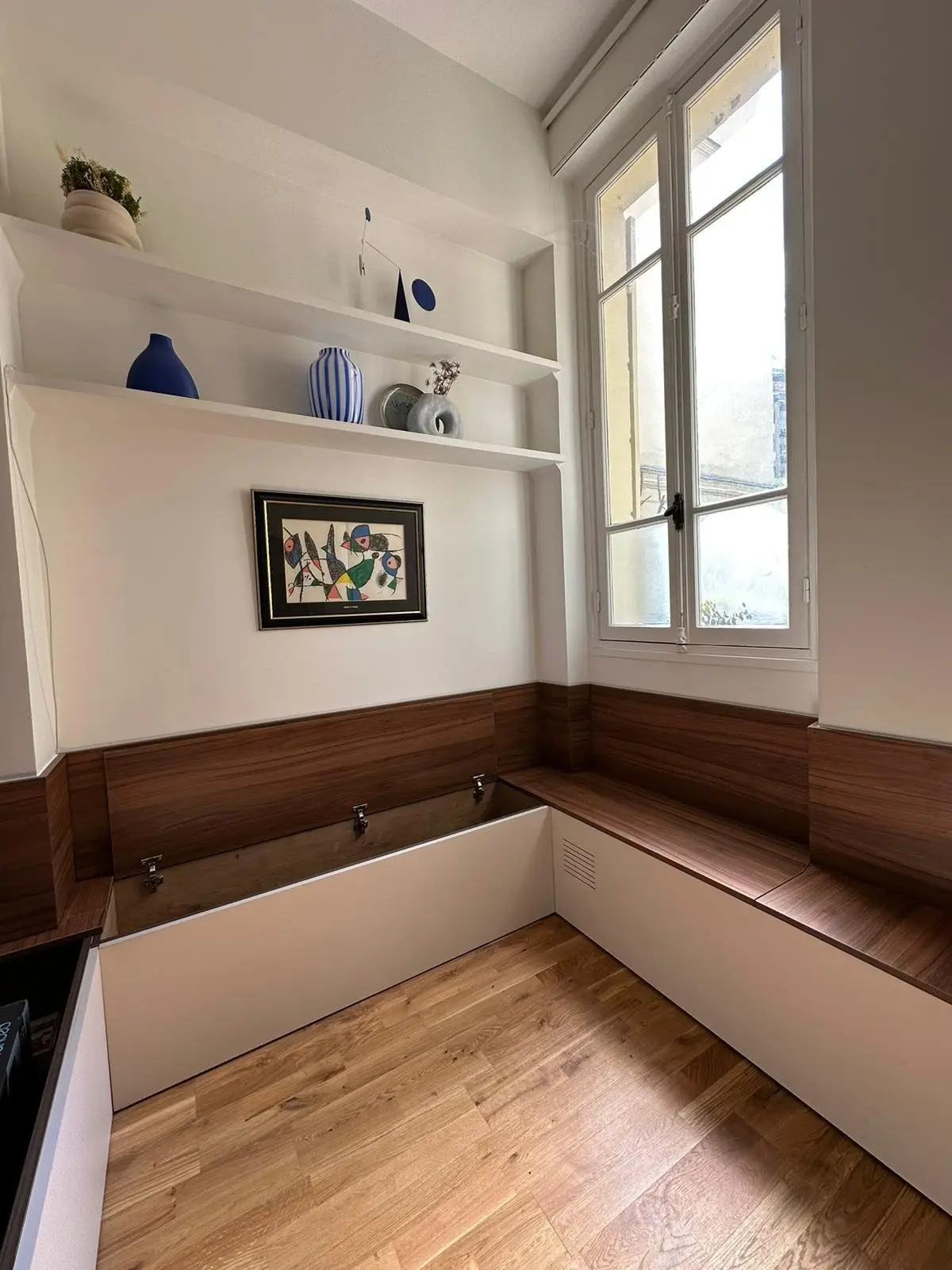 Banquette d’angle décor noyer avec coffres de rangement et dosseret bois, sous une fenêtre à petits bois