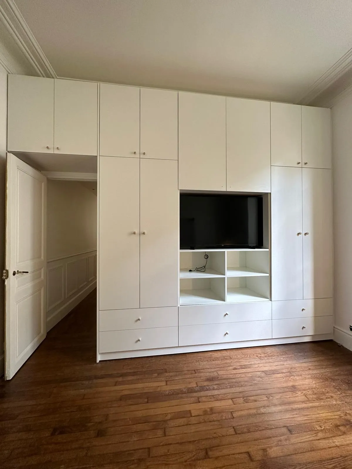 Placard blanc integrant un televiseur dans une niche centrale avec casiers et tiroirs, parquet ancien